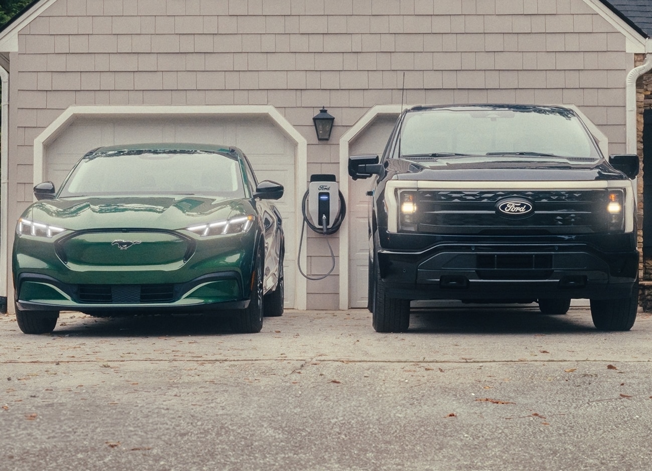 Two Ford electric vehicles are plugged into a home charger in a driveway in front of a home's garage.